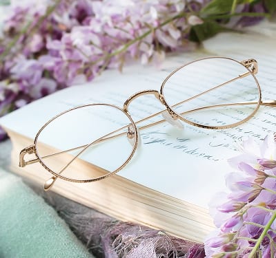 A pair of thin, Zenni gold-rimmed, oval glasses rests on an open book with lavender flowers